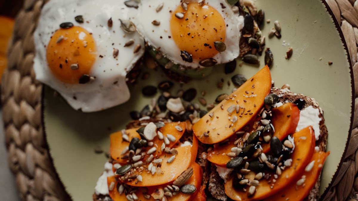 Delicious breakfast plate featuring avocado toast with fried eggs and persimmons, topped with seeds for a healthy start.